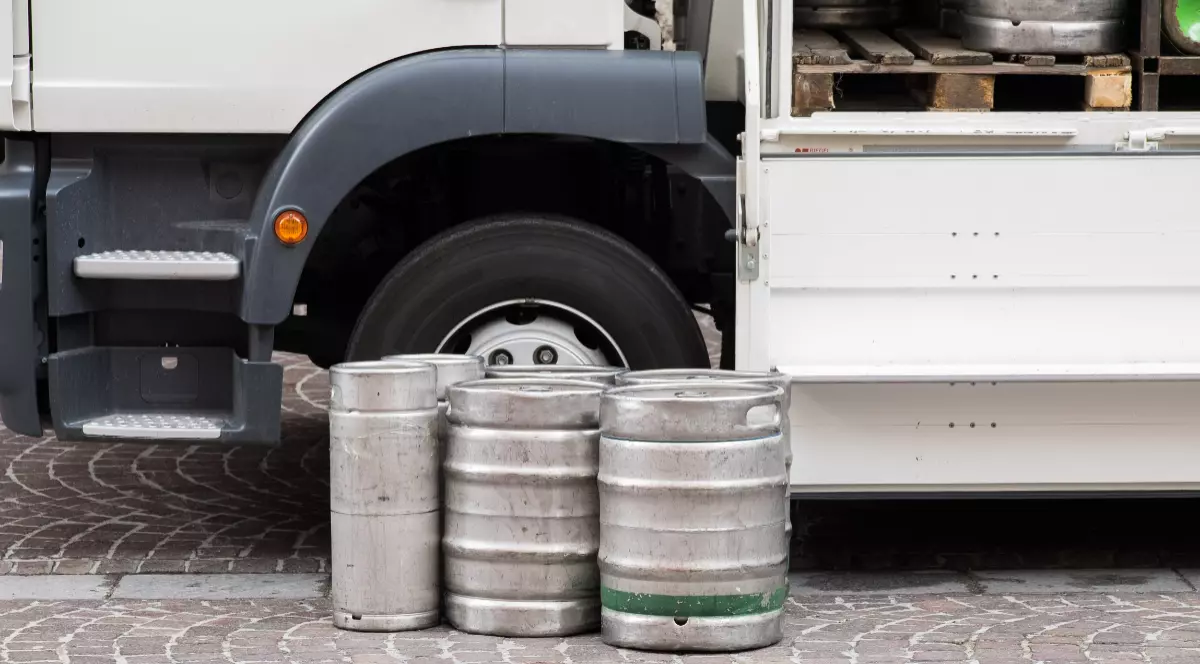Un grup de butoaie metalice de bere (keg-uri) așezate pe pavaj lângă o roată și remorca deschisă a unui camion alb de livrări.