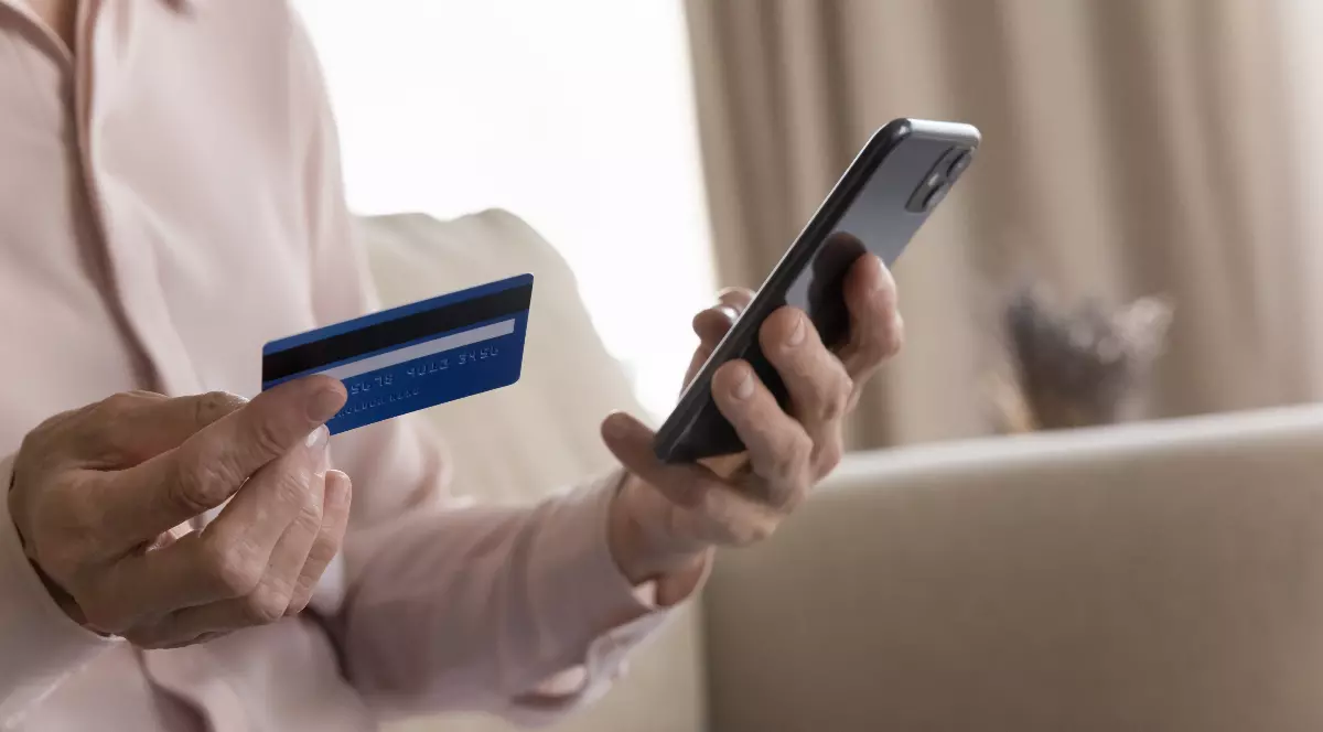 Imaginea prezintă o persoană care ține în mână un card bancar și un telefon mobil, sugerând efectuarea unei operațiuni financiare online. Fotografia este ilustrativă și face trimitere la utilizarea aplicațiilor de mobile banking și a plăților digita