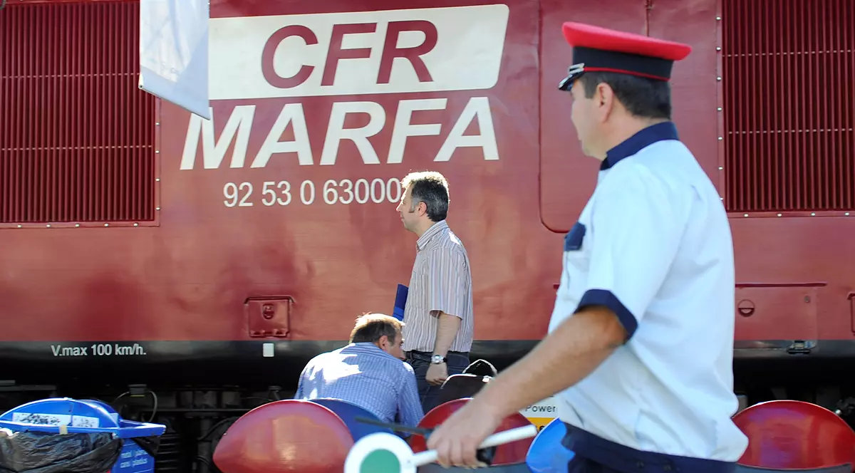Un vagon rosu pe care scrie CFR Marfa si oameni care trec prin fata lui