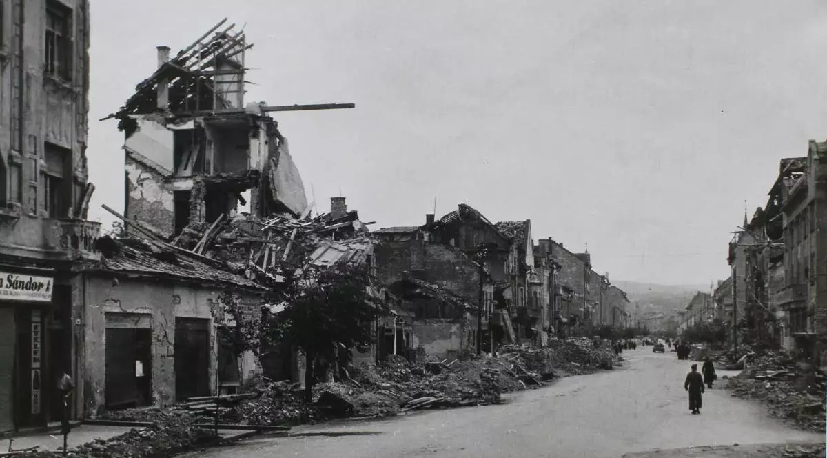 Fotografie alb-negru din Cluj, după bombardamentele americane din 2 iunie 1944. Mai multe clădiri de pe o stradă lungă sunt distruse aproape complet, cu pereți prăbușiți și mormane de dărâmături pe marginea drumului. În depărtare se văd câțiva oameni care merg printre ruinele orașului.