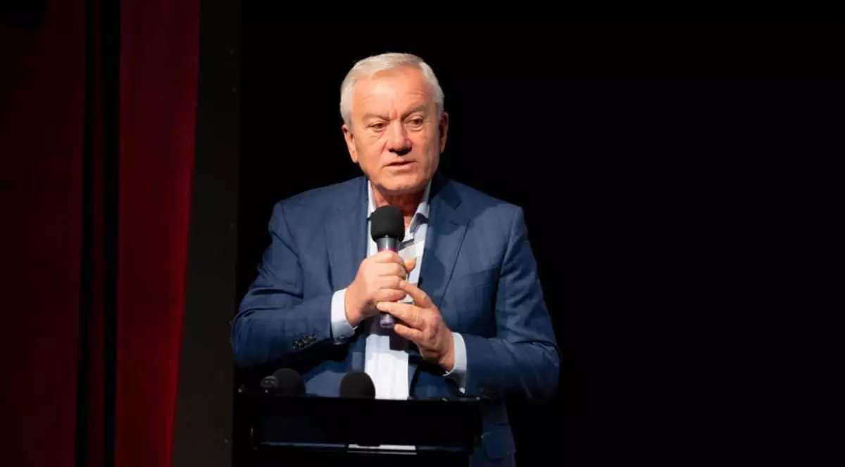 Constantin Toma, îmbrăcat în costum albastru, cămașă albă și cravată, cu microfonul în mână, pe fundal negru