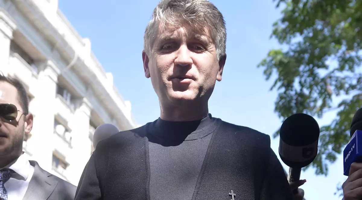 Cristian Pomohaci, îmbrăcat cu o bluză neagră și o vestă neagră, lângă o clădire, cu cerul albastru pe fundal