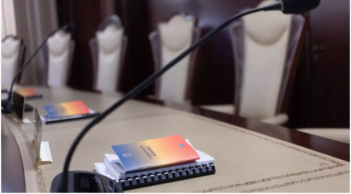 În imagine este prezentată o masă oficială de consiliu goală, având în prim-plan broșuri cu textul „Constituția României” și microfoane flexibile așezate în fața unui rând de scaune cu spătar înalt.