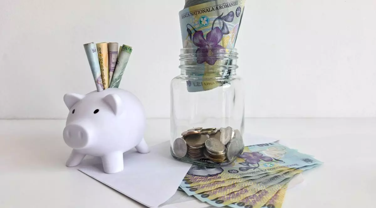 O pușculiță albă în formă de porc, plină de bani românești, sub ea este un plic alb umplut cu bancnote de 100 de lei, iar peste el este un borcan transparent din sticlă plin cu monede, iar deasupra hârtii de 100 de lei