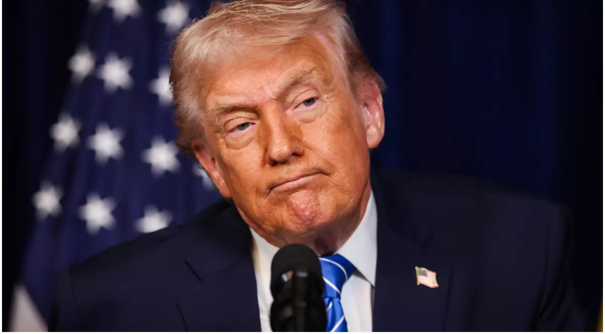 Portret în plan apropiat al lui Donald Trump având o expresie serioasă în fața unui microfon, cu steagul Statelor Unite ale Americii vizibil în fundalul albastru.
