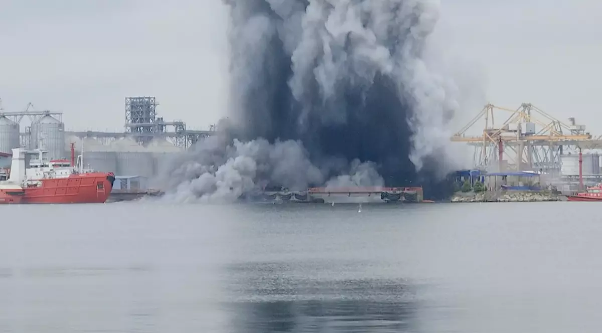 Imaginea surprinde momentul exploziei produse în zona portuară, unde un nor uriaș de fum negru și gri se ridică deasupra apei. În fundal se observă nave și instalații industriale specifice Portului Constanța, în timp ce coloana densă de fum domină întregul peisaj.
