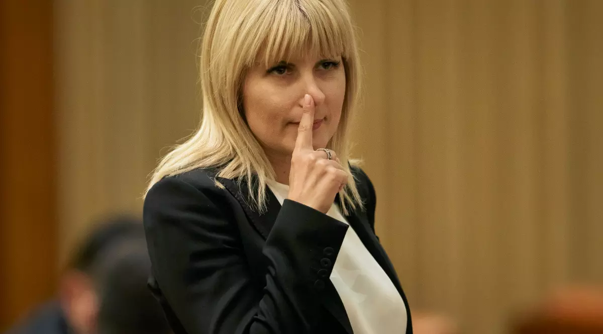 Imaginea o surprinde pe Elena Udrea într-un cadru oficial, îmbrăcată într-un sacou negru și o bluză albă, în timp ce își duce degetul la nas într-un gest care sugerează reflecție sau concentrare. Fotografia pare realizată în timpul unei ședințe sau al unui eveniment public, având un fundal discret și o atmosferă sobră.