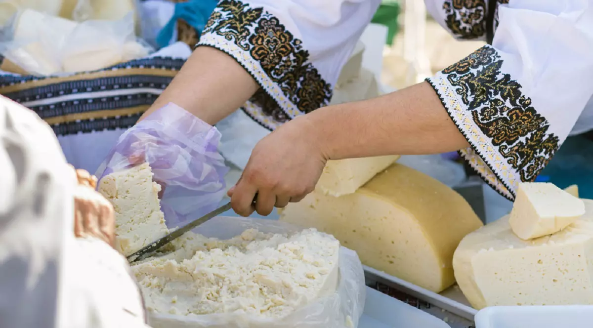 Care este diferenţa dintre brânză, urdă şi caş - Imagine cu o femeie îmbrăcată în costum popular care taie o bucată de brânză