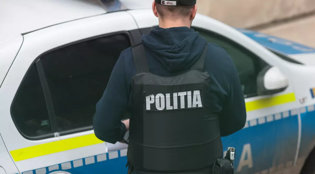 În imagine este prezentat din spate un bărbat ce poartă o vestă antiglonț inscripționată cu textul „POLIȚIA” și o șapcă neagră, stând lângă o autospecială de poliție alb-albastră.