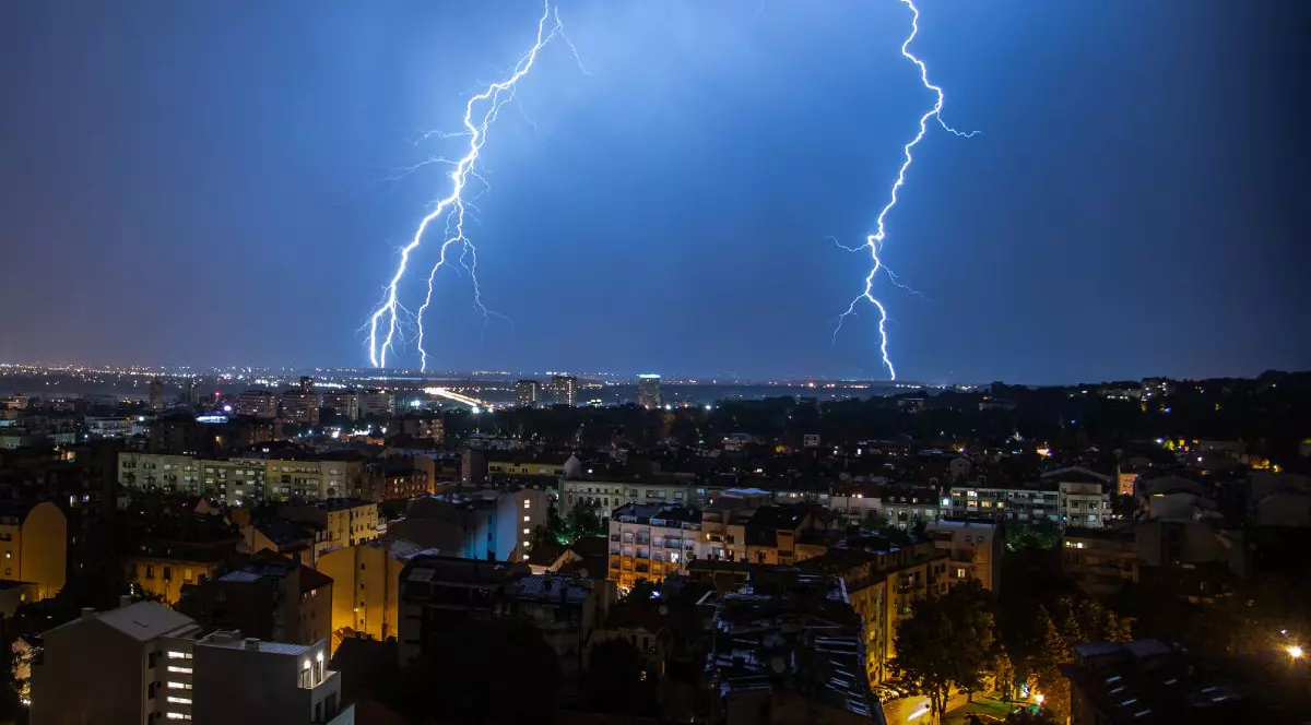 Furtună cu fulgere și cer înnorat peste un oraș, cu două fulgere care lovesc pământul în fundal