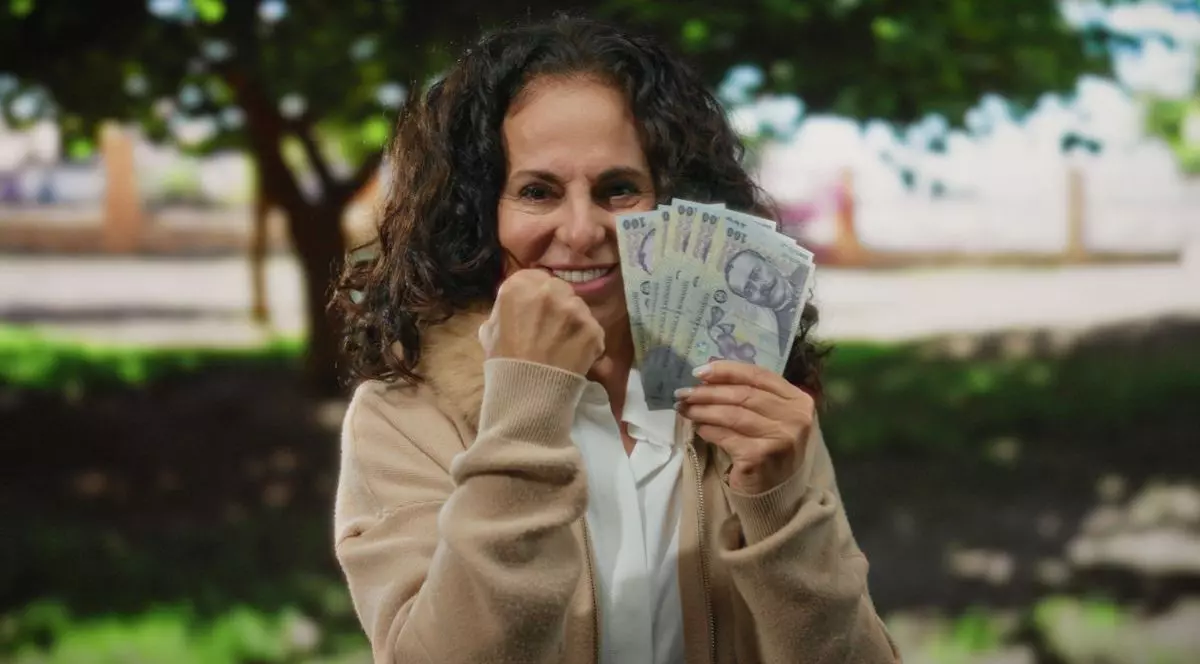 O femeie de vârstă mijlocie ține cu entuziasm în mână mai multe bancnote românești de 100 de lei, într-un parc, sugerând succes financiar