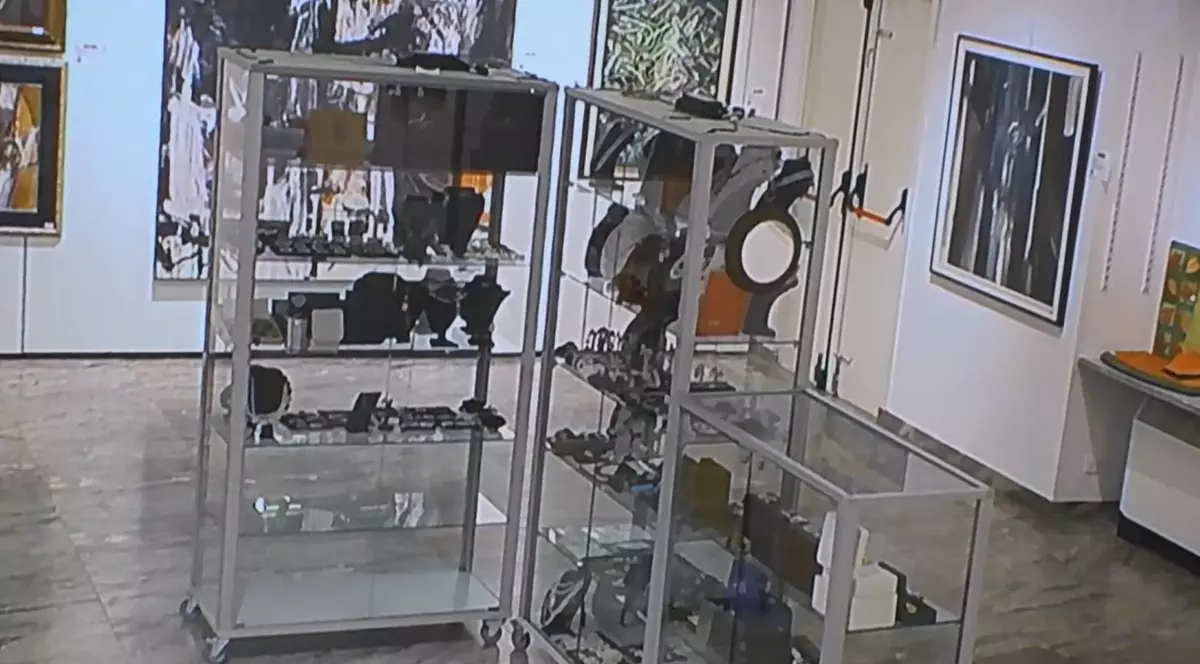 Interiorul unei galerii de artă sau case de licitații, unde se observă vitrine metalice cu geamuri transparente pline cu diverse obiecte decorative și bijuterii, în timp ce pereții din fundal sunt decorați cu numeroase tablouri și picturi abstracte.