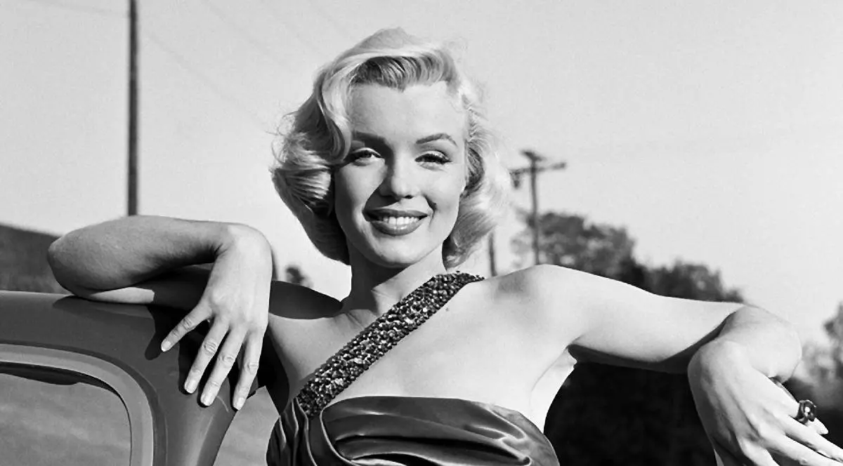 Actrița, modelul și cântăreața americană Marilyn Monroe (Norma Jeane Mortenson) stă sprijinită de o mașină sport și zâmbește în timp ce poartă rochia din filmul Cum să te căsătorești cu un milionar.