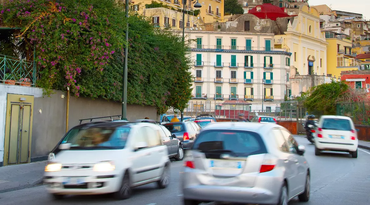 În imagine este prezentat traficul rutier aglomerat de pe o stradă urbană, cu mai multe mașini în mișcare ușor blurate, având pe fundal clădiri istorice în stil mediteranean specifice unui oraș din Italia.