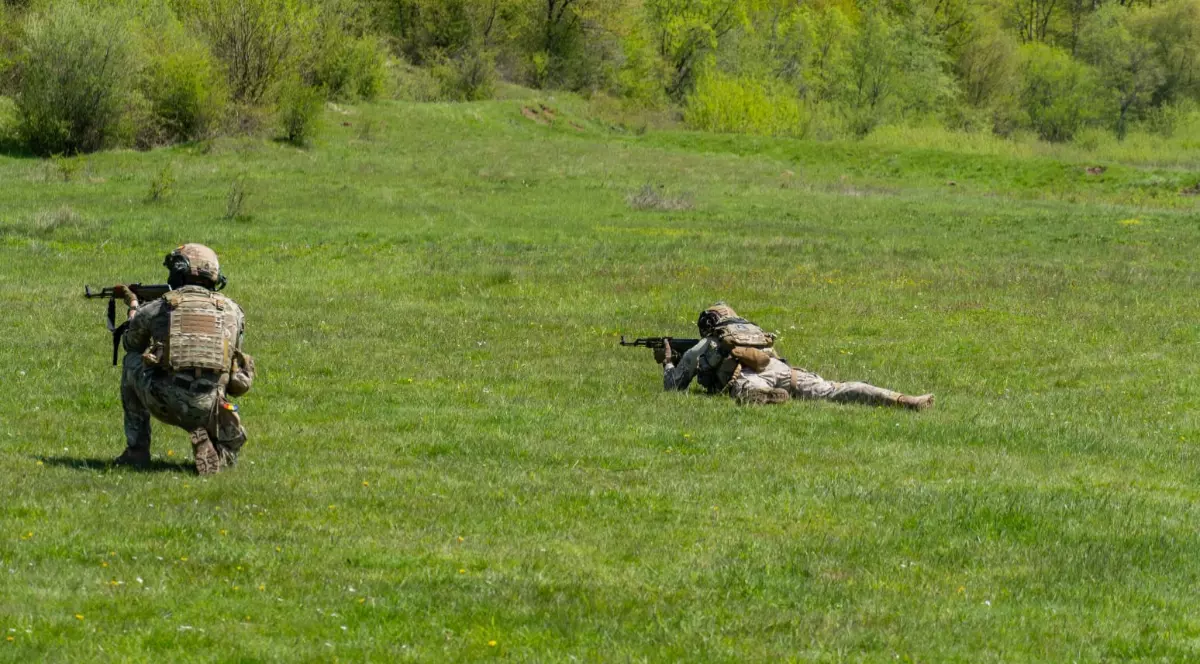 Doi soldați în echipament de camuflaj, în timpul unui exercițiu pe un câmp verde, unul fiind poziționat în genunchi, iar celălalt culcat la pământ, ambii țintind cu armele spre liziera pădurii din fundal.
