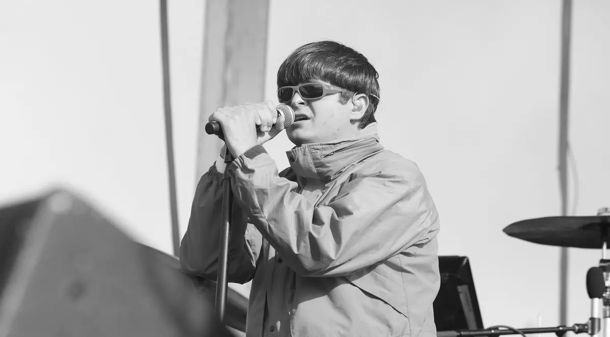 Imaginea îl surprinde pe un artist în timpul unui concert, cântând la microfon pe o scenă în aer liber. Acesta poartă o geacă largă și ochelari de soare, având o apariție distinctivă și o atitudine concentrată în timpul prestației.