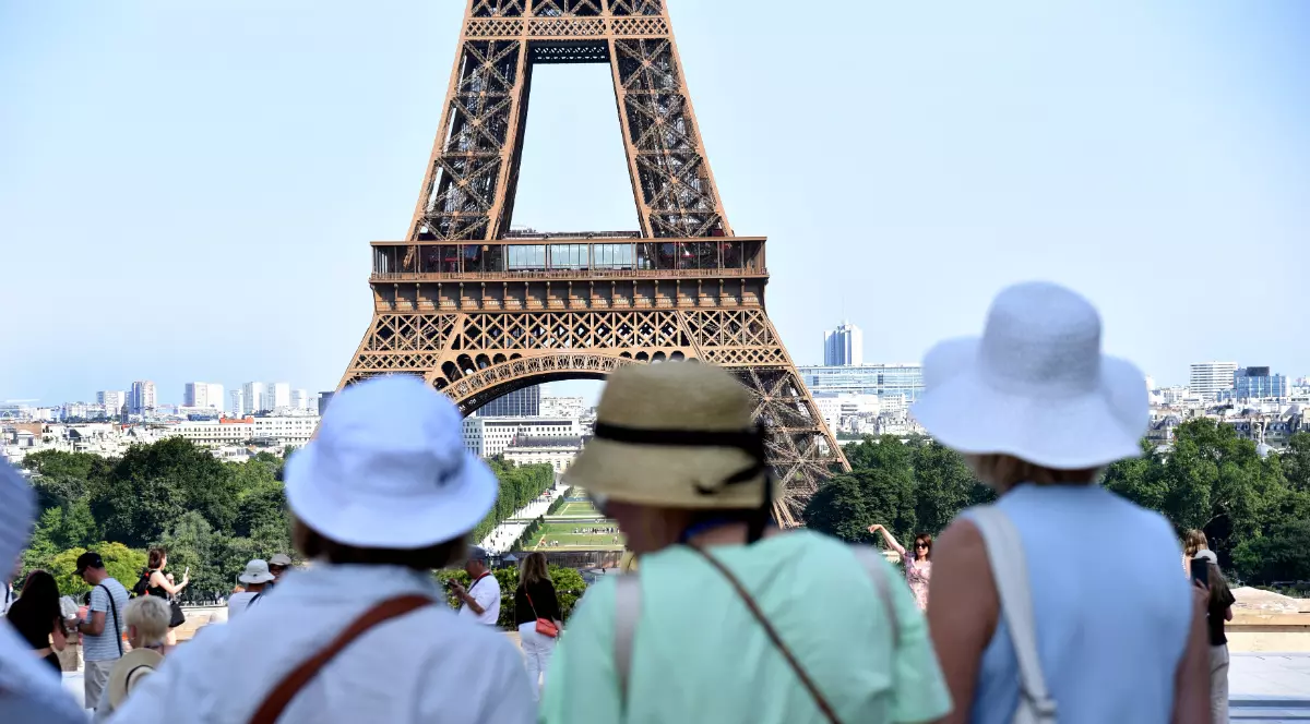 În prim-plan se află mai mulți turiști care admiră și fotografiază Turnul Eiffel, unul dintre cele mai cunoscute simboluri ale Parisului. Imaginea surprinde atmosfera unei zile însorite în capitala Franței, cu numeroși vizitatori adunați în zona de belvedere.