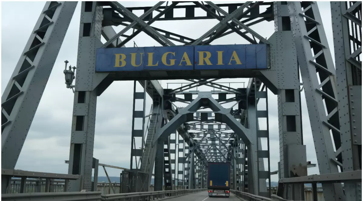 În imagine este un pod masiv cu structură metalică, pe care circulă un camion și deasupra căruia este montat un indicator albastru cu textul „BULGARIA”.