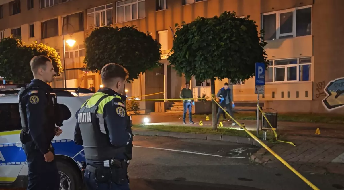 În imagine este prezentată o scenă nocturnă în care doi polițiști români în uniformă stau în prim-plan lângă o autospecială, în timp ce în fundal o zonă pietonală din fața unui bloc este securizată cu bandă galbenă de delimitare pentru investigații criminalistice.