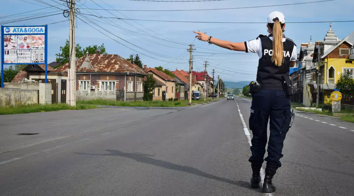 Polițistă de la Rutieră, îmbrăcată în uniformă, cu șapcă albă, pe mijlocul unei străzi din Cluj-Napoca, fotografiată din spate, cu mâna stângă întinsă, în timp ce oprește un șofer