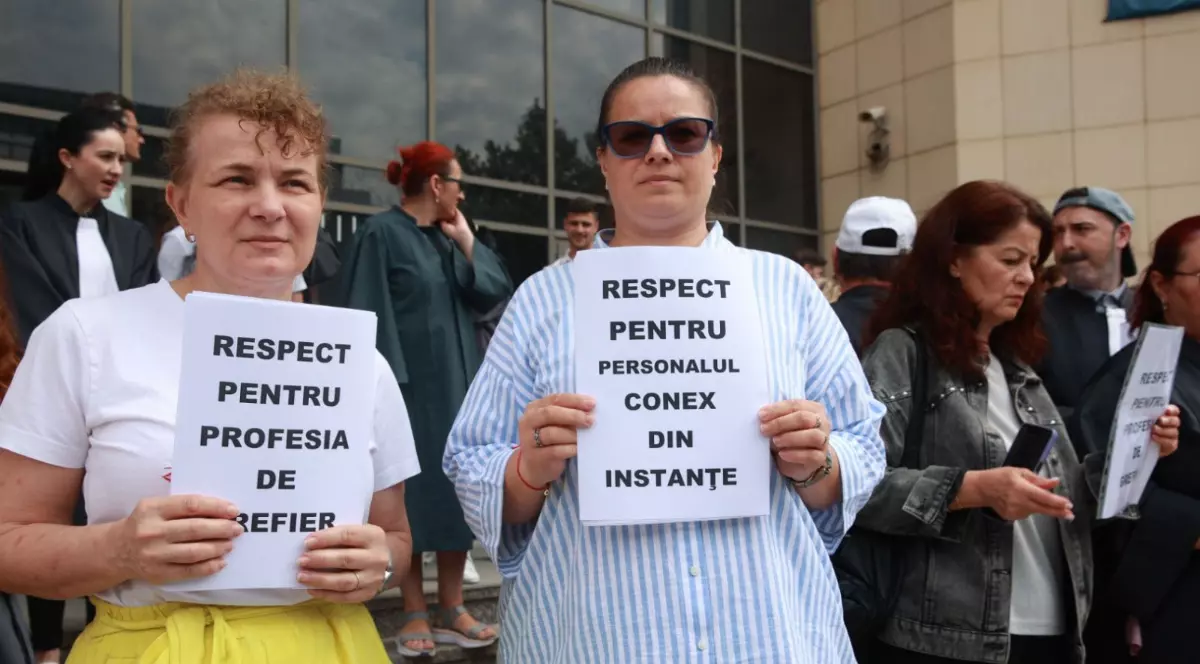 În imagine apar mai mulți participanți la un protest organizat în fața unei instituții publice, care afișează mesaje ce solicită respect pentru diferite categorii de personal din sistemul judiciar. Două femei din prim-plan țin pancarte cu mesajele „Respect pentru profesia de grefier” și „Respect pentru personalul conex din instanțe”, exprimând nemulțumirea față de situația actuală din domeniu.