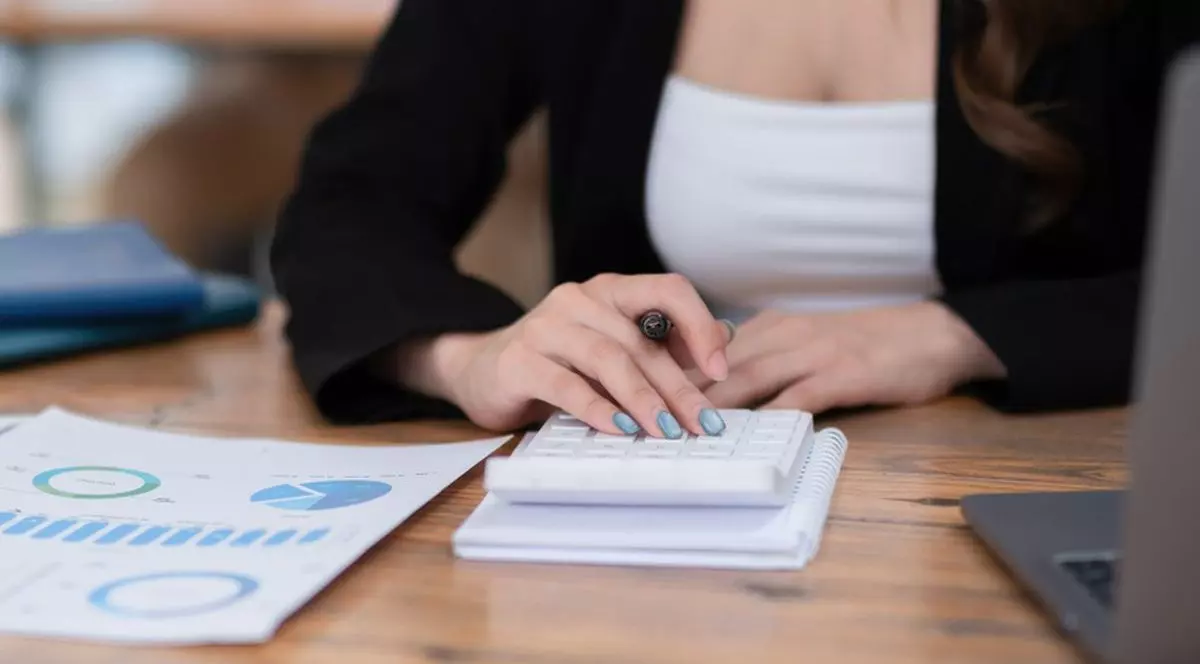 O contabilă cu maiou alb și sacrou negru stă la masă cu un calculator în față și calculează, în dreapta ei sunt niște hârtii cu grafice, iar în stânga este un laptoop