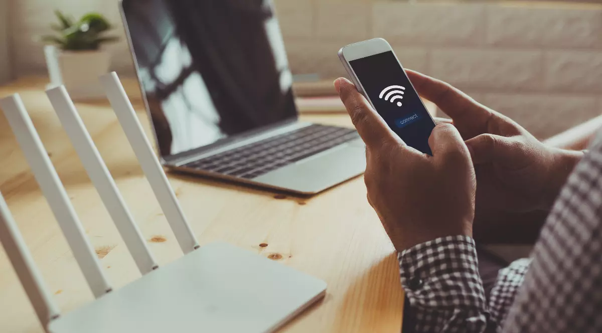 O persoană folosește un smartphone conectat la o rețea Wi-Fi, în timp ce pe birou se află un router wireless și un laptop.