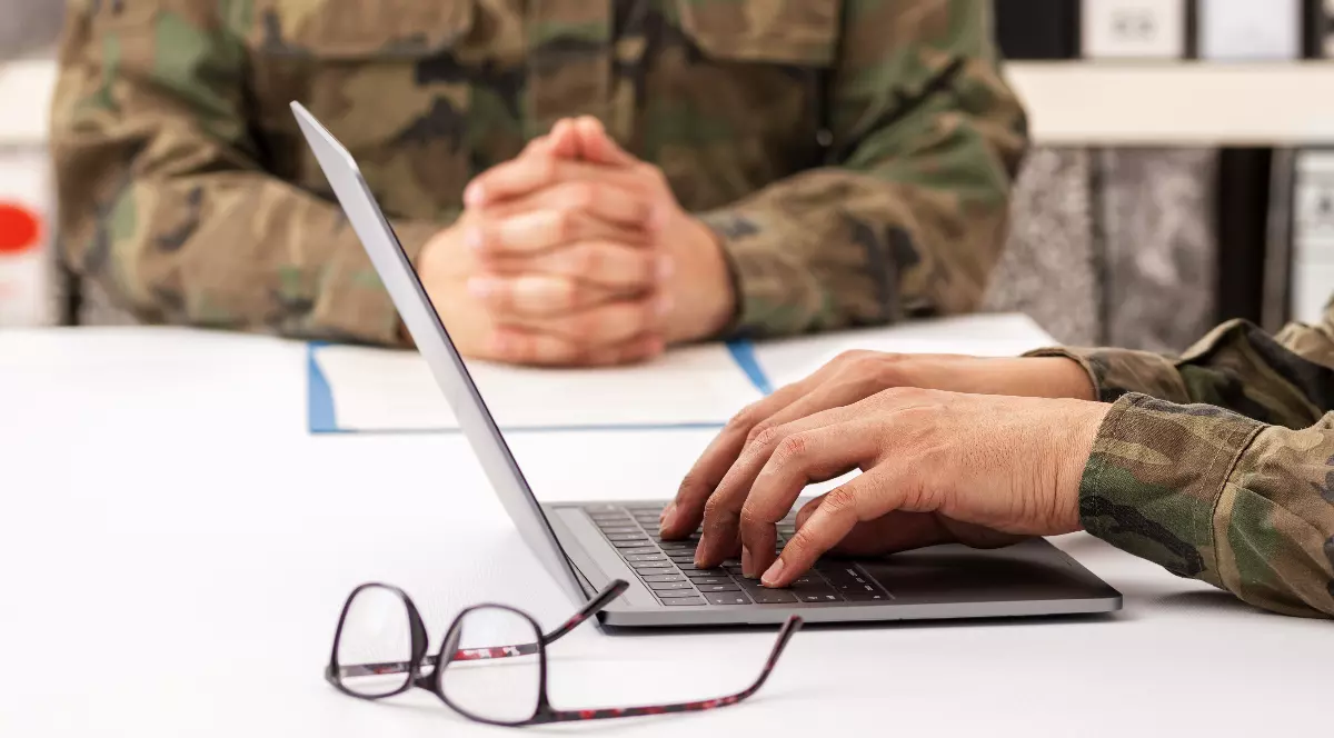 În imagine sunt prezentate în plan apropiat două persoane ce poartă uniforme militare de camuflaj așezate la o masă albă, una dintre ele tastând la un laptop lângă o pereche de ochelari, în timp ce cealaltă are mâinile împreunate în fundal.