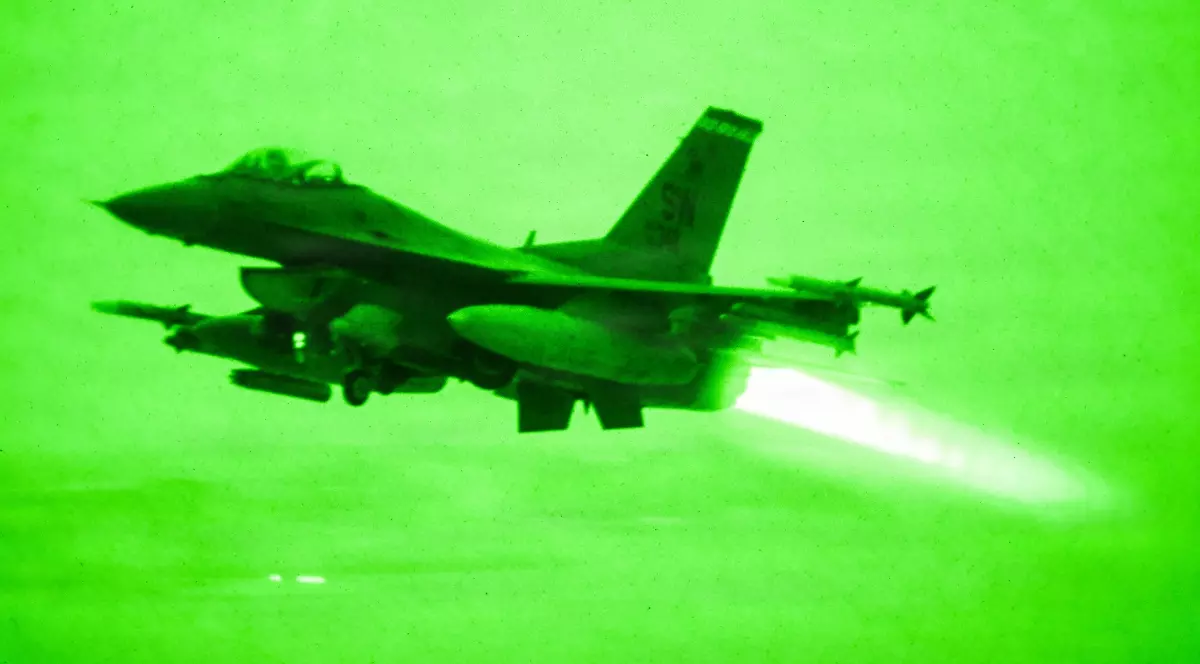 Un avion de vânătoare F-16 Fighting Falcon în zbor de noapte, capturat printr-un dispozitiv de vedere nocturnă în nuanțe intense de verde, în timp ce motorul său lasă o dâră puternică de lumină