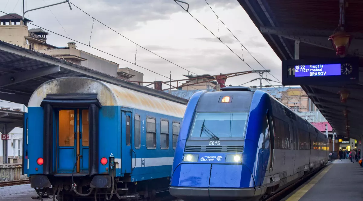 În imagine se află un tren modern al CFR Călători, staționat într-o gară alături de un vagon clasic al companiei. Contrastul dintre cele două garnituri evidențiază diferențele de design și tehnologie dintre materialul rulant nou și cel aflat de mai mulți ani în exploatare.