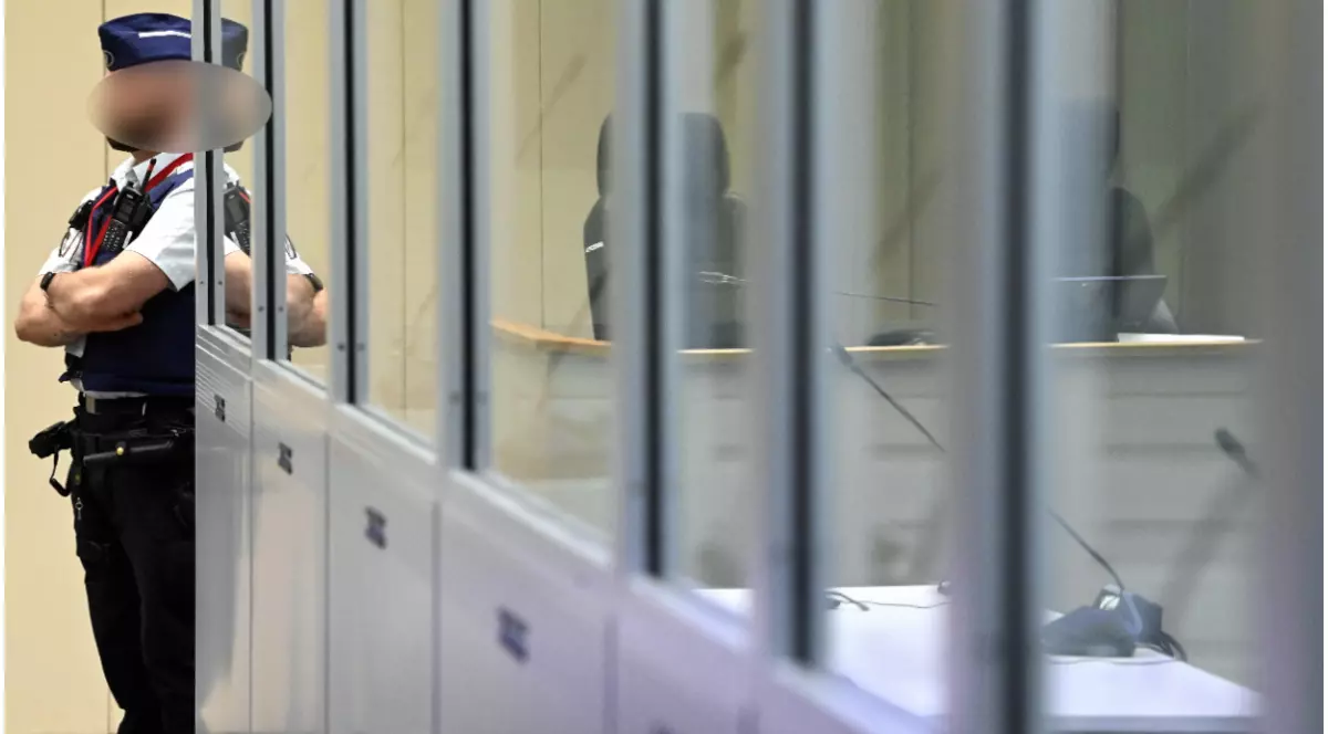 Un ofițer de poliție cu brațele încrucișate și fața blurată stând de pază în stânga unei cabine securizate cu geamuri transparente, în interiorul unei săli de judecată.