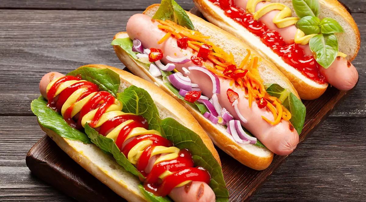 Trei hot-dogi cu sosuri diferite așezate pe masă.