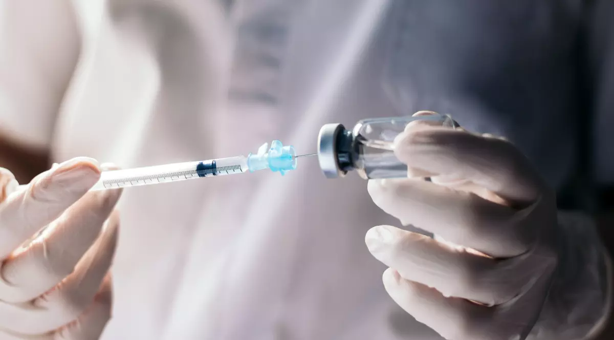 Un cercetător îmbrăcat în echipament medical de protecție pregătește o doză de vaccin, extrăgând lichidul dintr-un flacon cu ajutorul unei seringi. Imaginea sugerează un context de laborator sau de testare clinică, asociat dezvoltării și administrării vaccinurilor.
