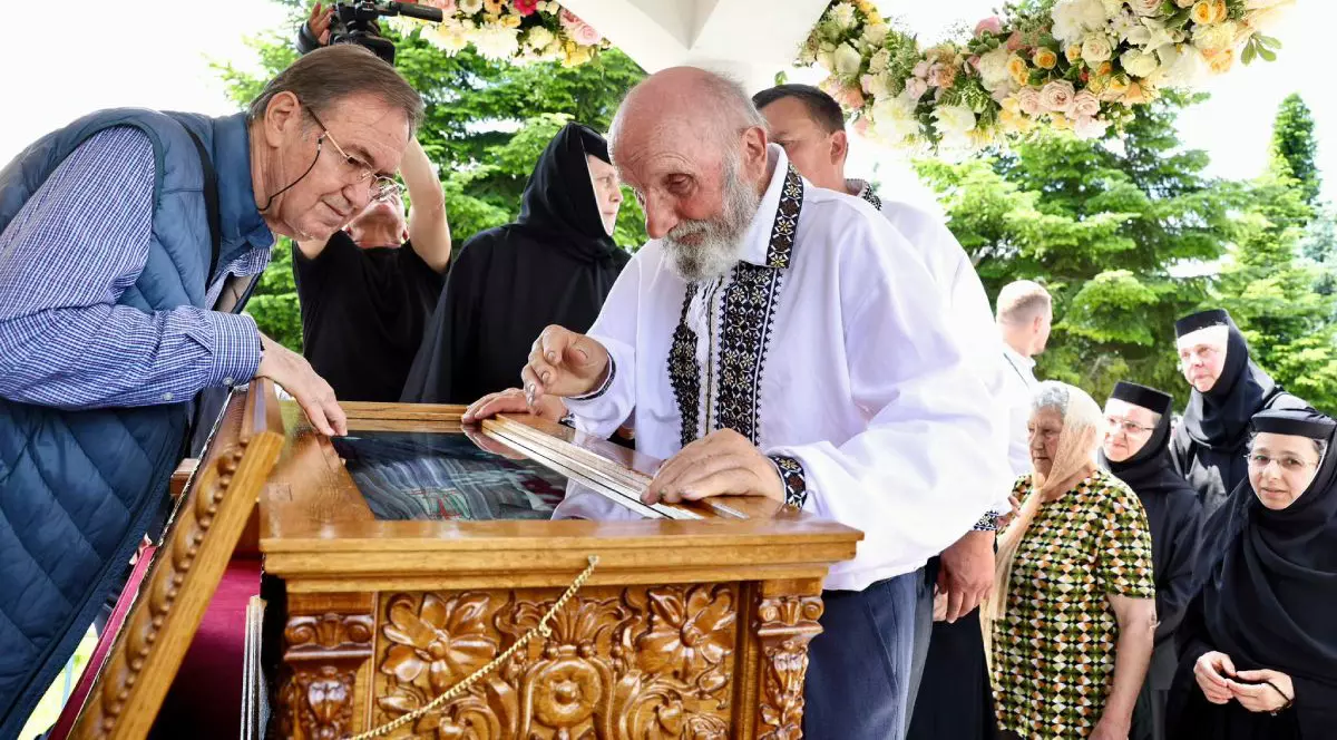 Vasile Lazăr, tatăl Sfintei Elisabeta de la Pasărea, îmbrăcat cu o ie românească, lângă racla cu moaștele fiicei sale, Sfânta Elisabeta de la Pasărea și alți oameni pe fundal