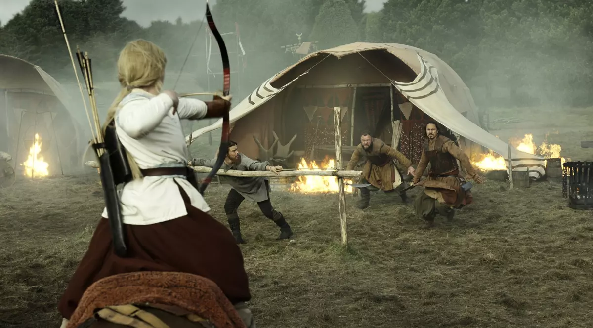 Imaginea surprinde o scenă tensionată de luptă într-o tabără medievală, unde o femeie călare țintește cu arcul spre mai mulți adversari care se apropie. În fundal se văd corturi, flăcări și fum, elemente care accentuează atmosfera dramatică și pericolul iminent