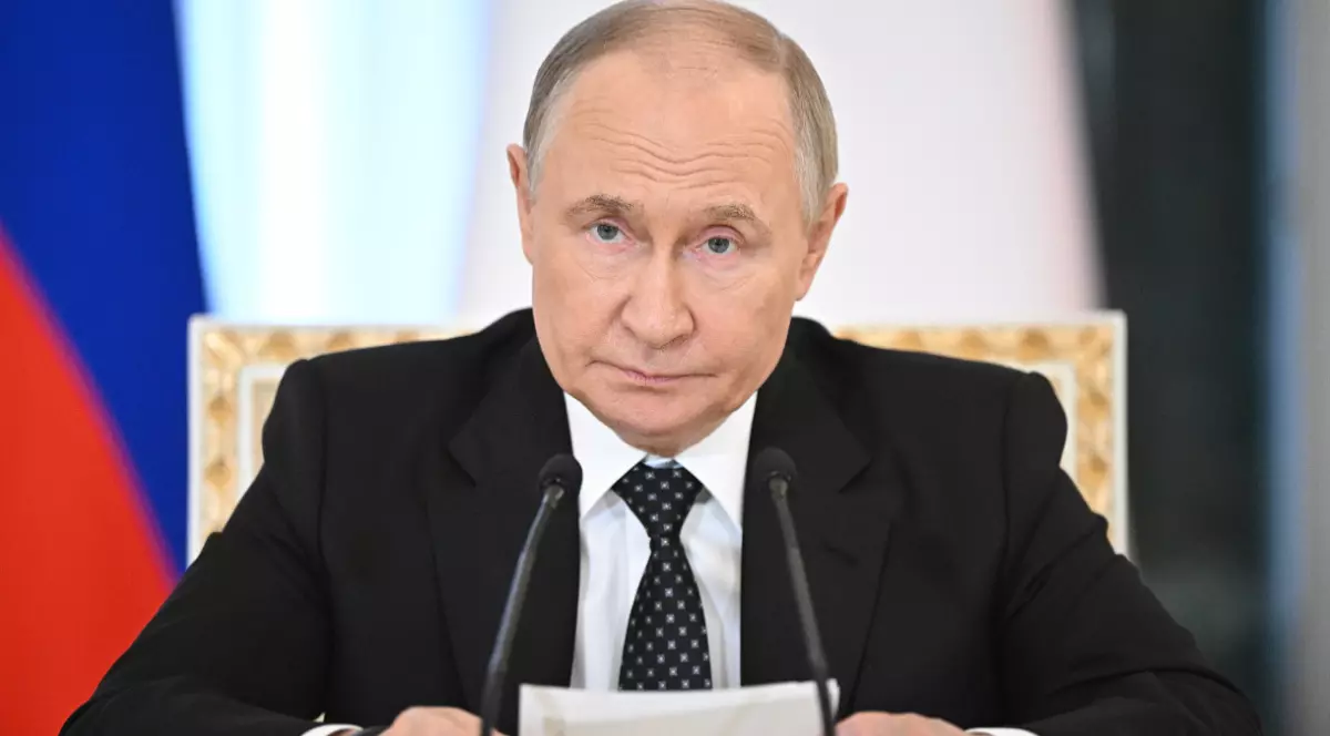Președintele rus Vladimir Putin, îmbrăcat în costum negru, cămașă albă și cravată cu buline albe așezat la o masă în fața a două microfoane și ținând niște foi în mână, având steagul Rusiei parțial vizibil în fundal