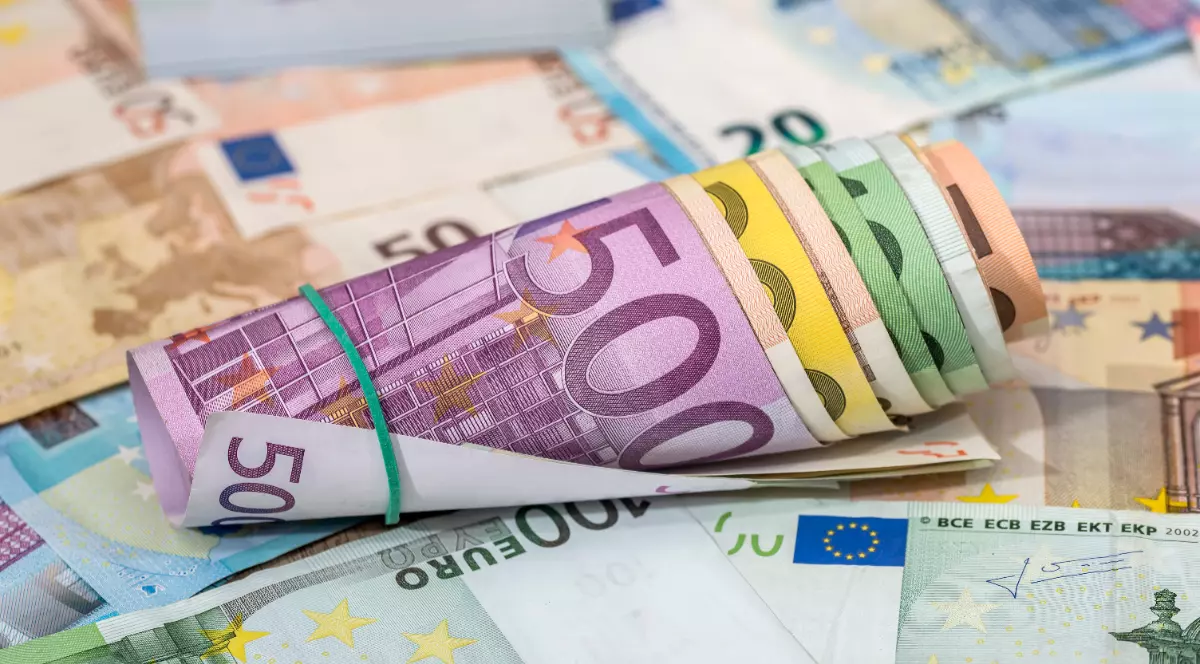Imaginea prezintă un sul de bancnote euro cu valori mari legat cu un elastic de cauciuc, așezat pe un fundal format din alte bancnote împrăștiate.