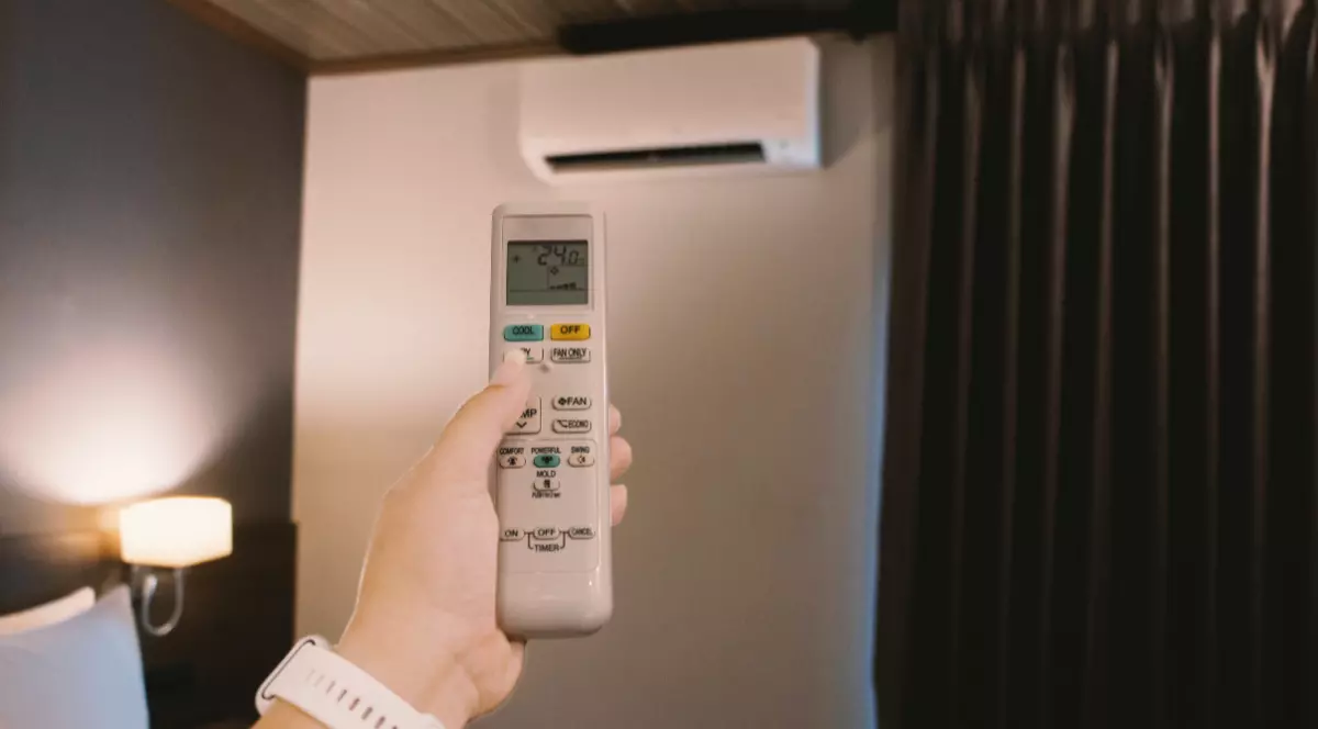 O femeie care ține în mână telecomanda unui aparat de aer condiționat, acesta este montat în peretele de deasupra patului și este pornit