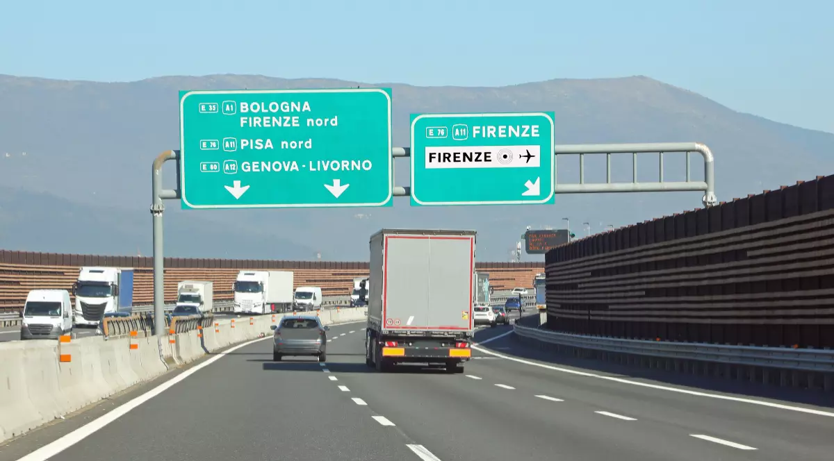 Imaginea prezintă traficul de pe o autostradă din Italia, cu vehicule aflate în mers sub două panouri verzi mari de semnalizare care indică direcțiile către orașe precum Bologna, Pisa și Firenze.