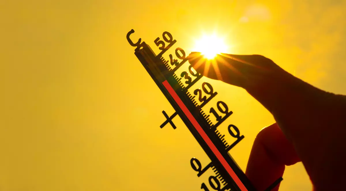 Imaginea prezintă o mână care ține un termometru orientat spre soarele puternic, indicând o temperatură ridicată de peste 35°C pe un fundal galben strălucitor.