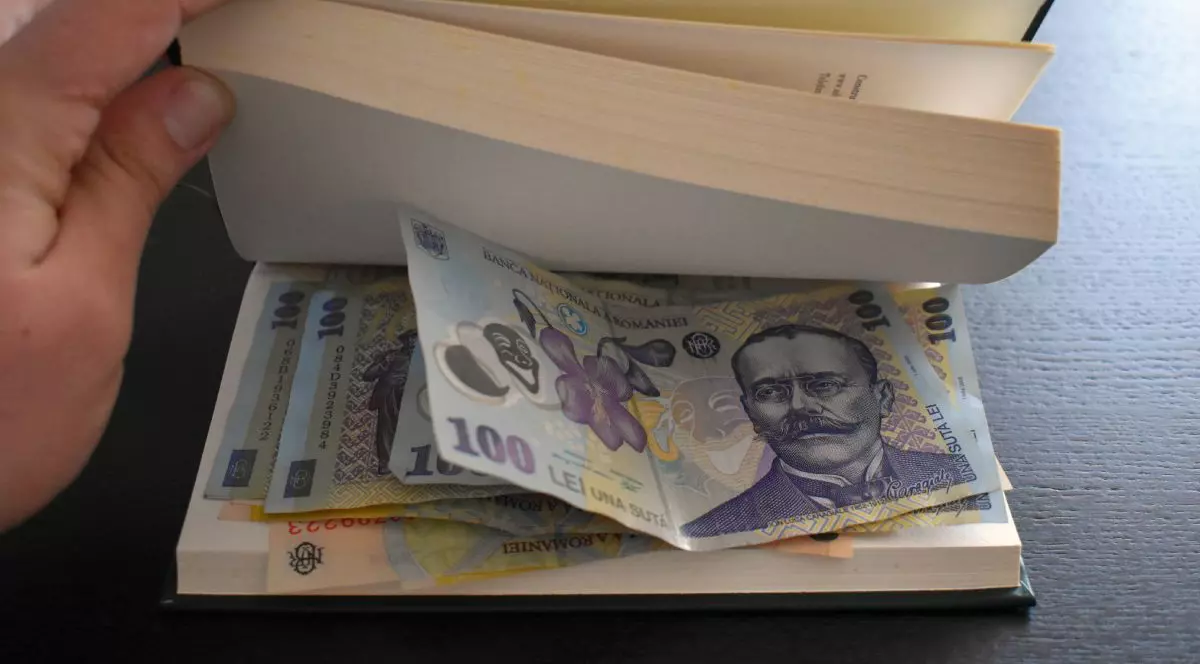Bani, în bancnote de 100 de lei, ascunși într-o carte cu pagini albe, așezată pe o masă de culoare neagră