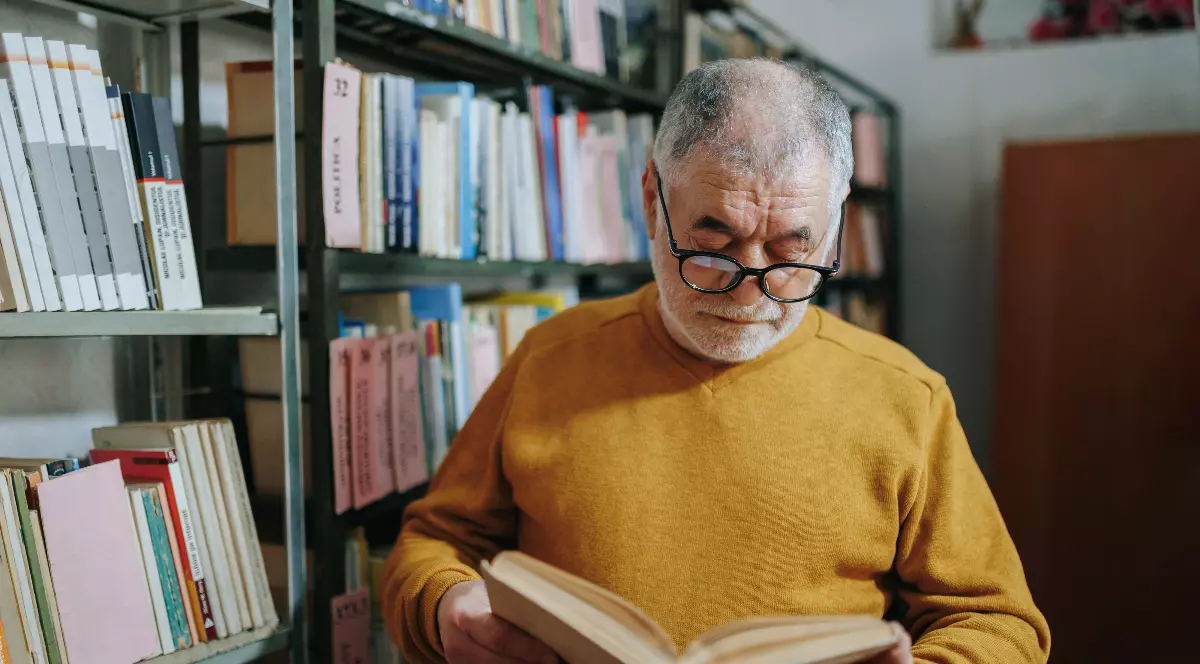 Un bărbat răsfoiește o carte într-o bibliotecă, fiind înconjurat de rafturi pline cu volume. Pare concentrat și atent, în timp ce citește cu interes conținutul cărții.