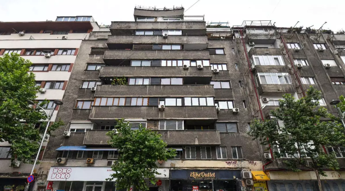 Imaginea prezintă fațada cenușie a unui bloc vechi de locuințe cu mai multe etaje și magazine la parter, situat într-o zonă urbană.
