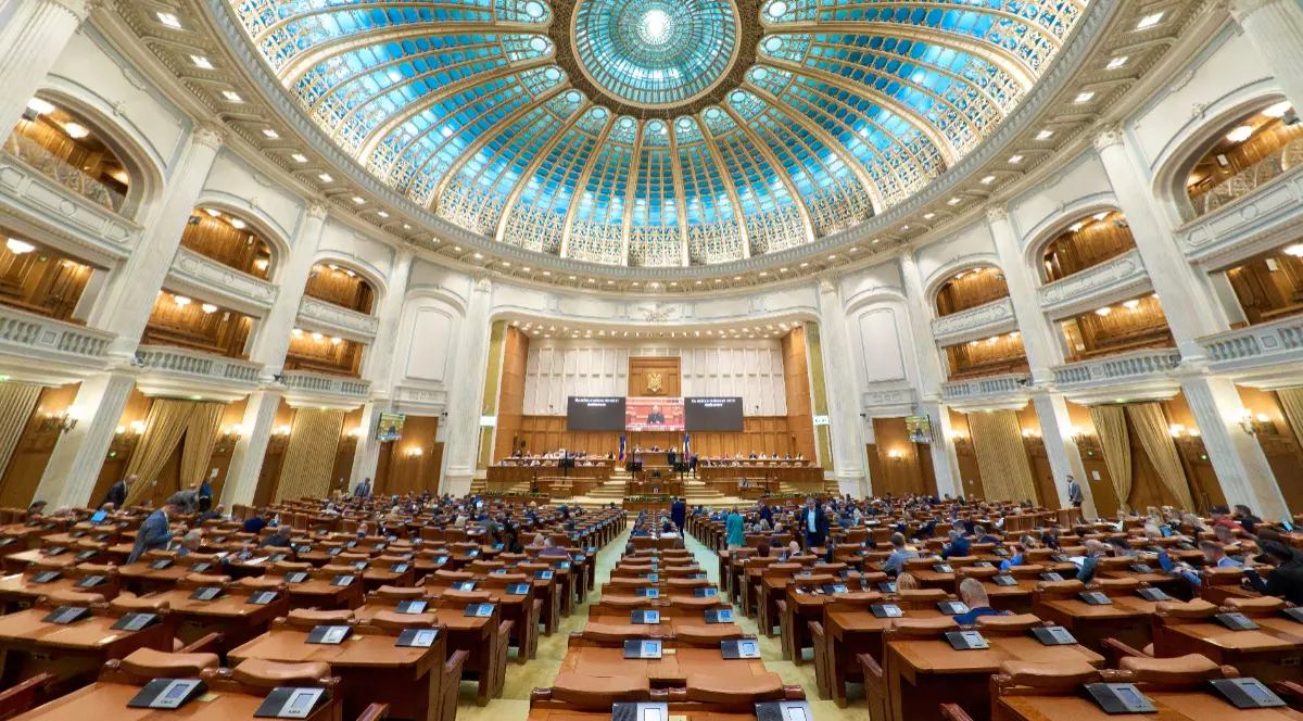 Imaginea prezintă o vedere de ansamblu din interiorul plenului Camerei Deputaților din Palatul Parlamentului, cu un vitraliu albastru pe cupolă și băncile parlamentarilor pregătite pentru ședință.