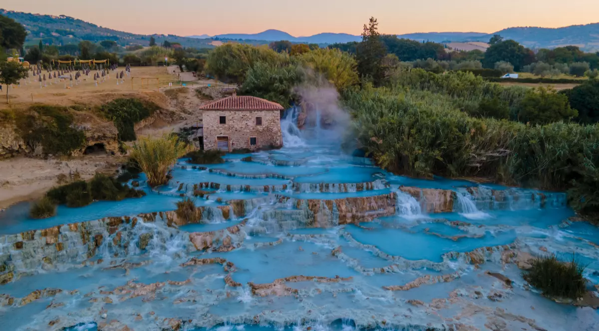 Vedere aeriană u Cascate del Mulino din Saturnia, Toscana. Cascade termale naturale cu bazine din travertin pline cu apă turcoaz, aburi calzi și o moară veche din piatră în fundal.