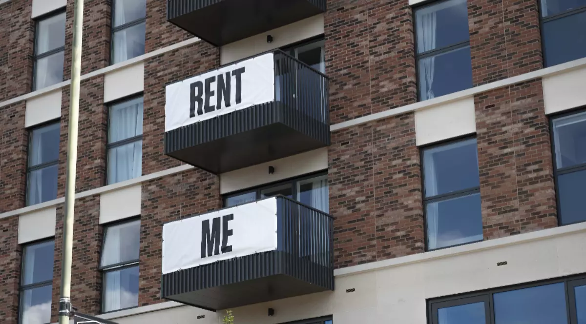 Imaginea prezintă fațada unei clădiri din cărămidă, ale cărei balcoane au afișate bannere albe ce formează textul „RENT ME” (Închiriază-mă).