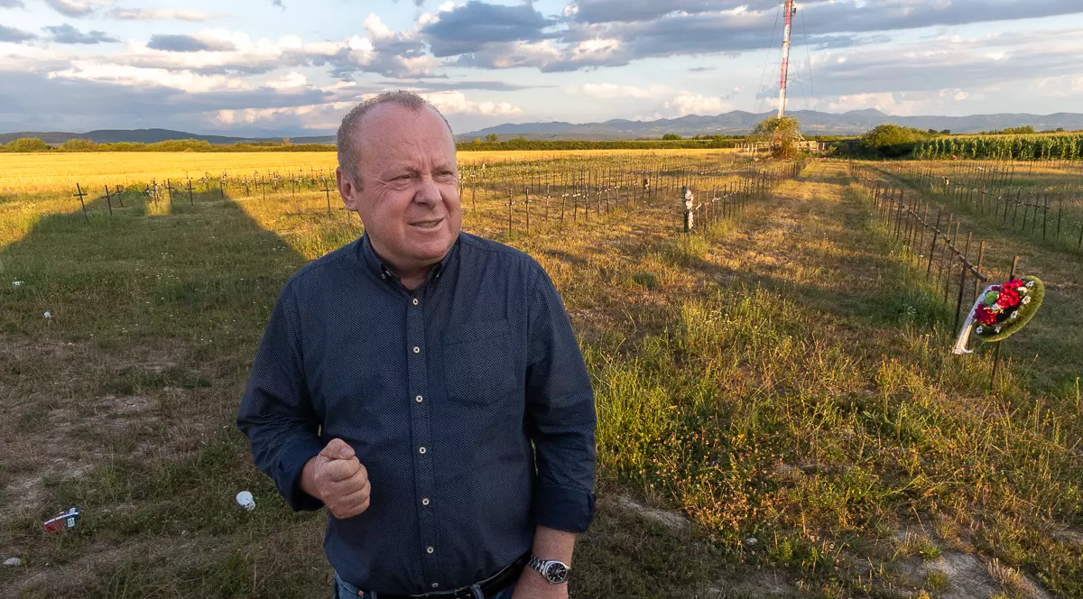 Un barbat in camasa de culoare inchisa sta cu pumnii stransi in fata unui cimitir cu multe cruci