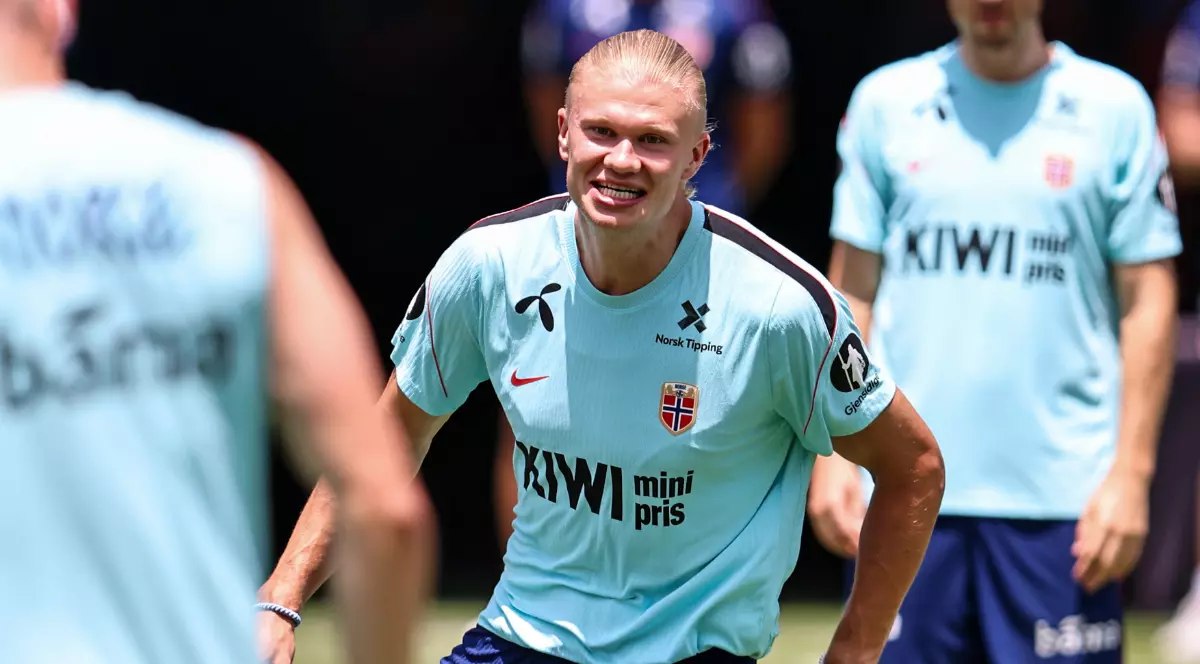 Fotbalistul din prim-plan participă la un antrenament al naționalei Norvegiei, fiind surprins într-un moment de intensitate și concentrare. În jurul său se află colegii de echipă, pe teren, în timpul unei ședințe de pregătire desfășurate pe vreme însorită.