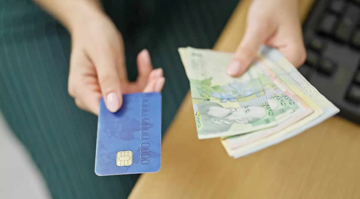 O femeie care stă la un birou de lemn maro și ține în mână un card de credit albastru și mai multe hârtii de 1 lei, 10 lei și 100 de lei