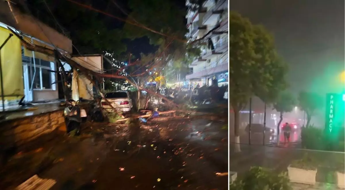 colaj format din două fotografii de noapte care surprind efectele unei furtuni puternice într-o zonă urbană, reprezentând orașul Sarande. Fotografia din partea stângă prezintă o stradă unde un copac masiv a fost doborât, trunchiul și crengile acestuia prăbușindu-se peste un autoturism și un scuter parcate lângă un trotuar pietruit. Asfaltul ud este acoperit de numeroase resturi vegetale și frunze rupte, iar pe fundal se observă clădiri cu mai multe etaje, balcoane și vitrine luminate ale unor spații comerciale. Fotografia din partea dreaptă ilustrează o altă porțiune de stradă în timpul unei ploi torențiale care reduce semnificativ vizibilitatea, creând un efect de ceață deasă. Pe marginea trotuarului umed și puternic reflexiv iese în evidență o firmă luminoasă verticală cu inscripția „PHARMACY”. În depărtare, printre copacii de pe aliniamentul stradal, se conturează silueta unei persoane care se deplasează prin precipitații, alături de luminile estompate ale unor mașini aflate pe șosea.